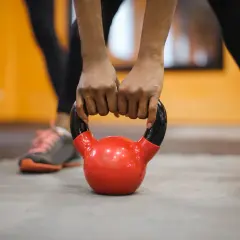 Dłonie zaciskają uchwyt czerwonej kettlebell, gotowe do treningu crossfit. W tle widać inne osoby ćwiczące.