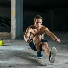 Mężczyzna wykonuje pistol squat w opuszczonym budynku. Dwa żółte kettlebelle leżą na ziemi.