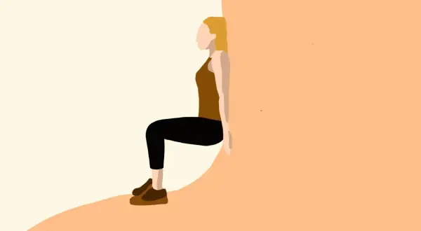 Kobieta w brązowym topie i czarnych legginsach wykonuje ćwiczenie wall sit, opierając plecy o ścianę.