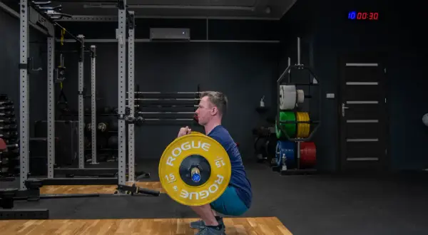 Mężczyzna wykonuje zercher squat z ciężarem w zgięciu łokci. Siłownia z wolnymi ciężarami i sprzętem do treningu siłowego.