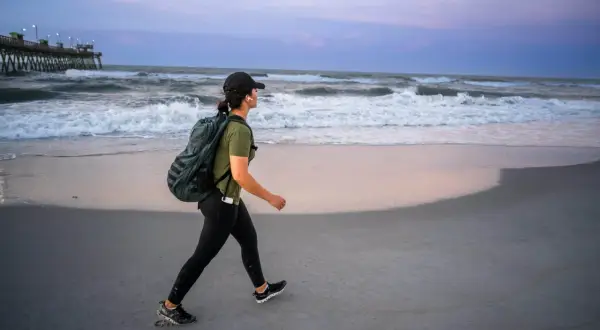 Kobieta z plecakiem na plaży, ćwicząca rucking przy falach oceanu.