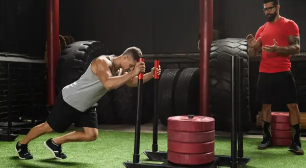 Mężczyzna wykonuje ćwiczenie sled push, pchając obciążony sprzęt. Trener obserwuje jego wysiłek.