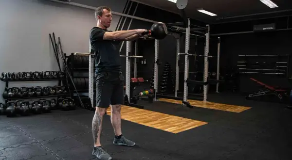 Mężczyzna ćwiczy z kettlebell w siłowni. W tle stojaki z ciężarami i sprzęt do treningu siłowego.