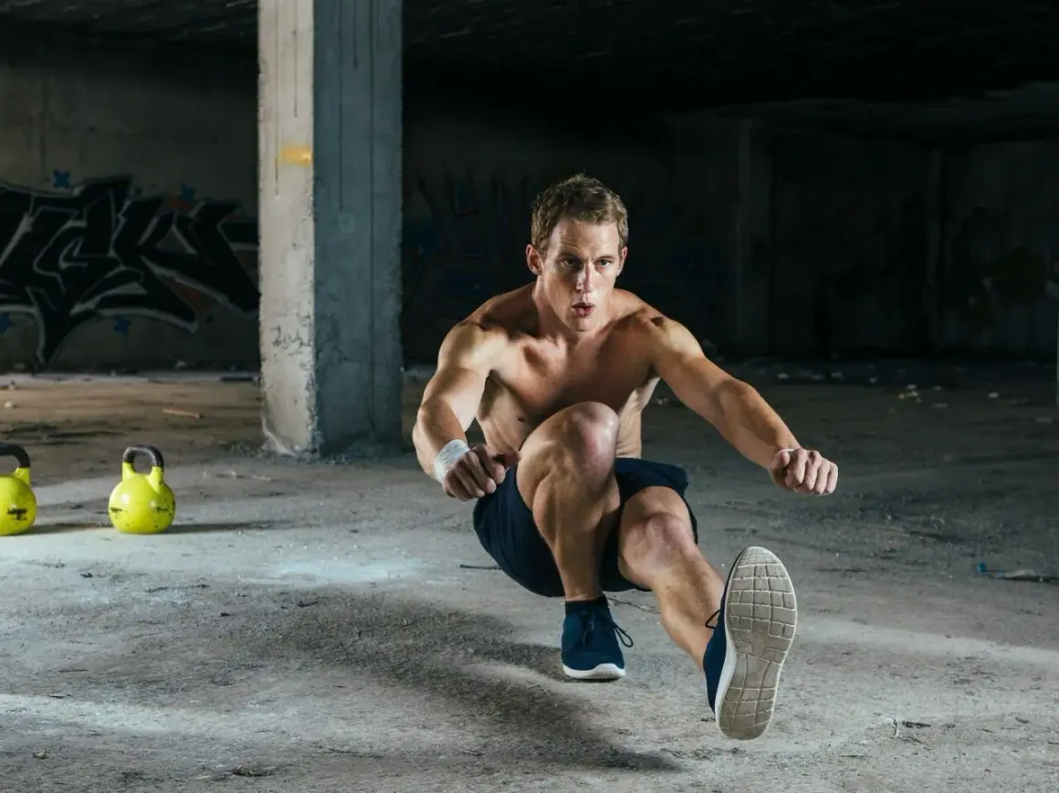Mężczyzna wykonuje pistol squat w opuszczonym budynku. Dwa żółte kettlebelle leżą na ziemi.