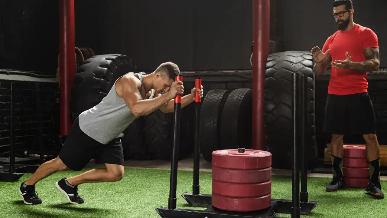 Trener motywuje zawodnika podczas intensywnego ćwiczenia sled push na siłowni.
